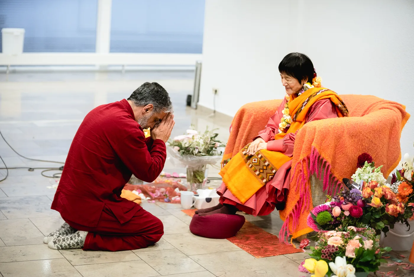 Ein Mann kniet mit gefalteten Händen vor Yogmata Keiko Aikawa, die von Blumen umgeben sitzt