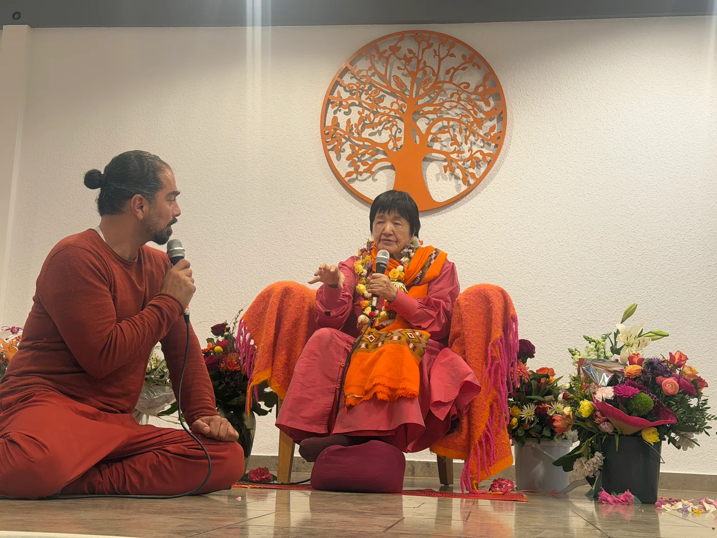 Yogmata Keiko Aikawa sitzt mit Mikrofon und spricht, neben ihr ein Übersetzer, dahinter ein Lebensbaum an der Wand