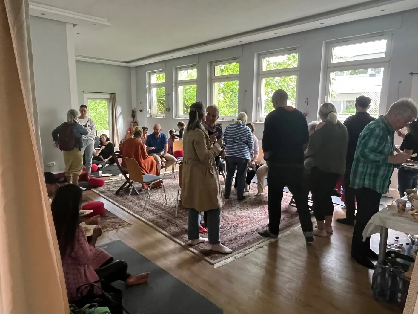Menschen stehen und sitzen nach der Meditation in Gruppen im hellen Raum, mit Tee und Tellern im Gespräch